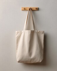 Plain natural canvas tote bag hanging from a simple wooden hook on a neutral white wall.
