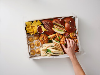 A hand reaches for a delightful variety of snacks displayed on a platter, featuring sandwiches, pastries, chips, and desserts. Perfect for parties or casual get-togethers