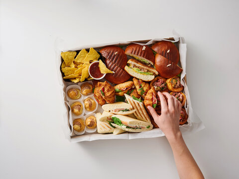 A hand reaches for a delightful variety of snacks displayed on a platter, featuring sandwiches, pastries, chips, and desserts. Perfect for parties or casual get-togethers