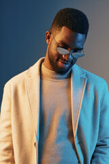 Stylish African male model in a light coat and sunglasses, showcasing a thoughtful expression against a blue background, highlighting modern fashion trends and elegance