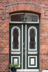 old brick wall, Above the doors, a large transom window with stained or textured glass is set beneath a brick archway. An old-fashioned lantern hangs above the door, and a small potted plant 