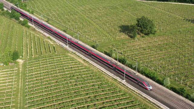 A red high-speed train races across vineyard-covered hills, blending modern transportation with nature. Captured from above.