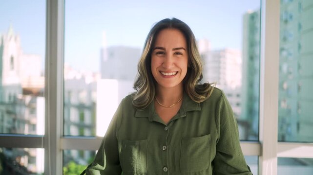 Successful Latin businesswoman in green shirt standing at modern desk with laptop against floor-to-ceiling windows with bright urban skyline