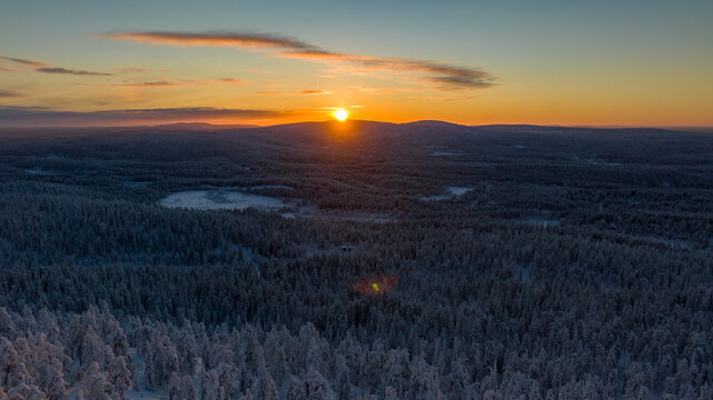 Aerial view of the sun rising over snow-covered forests and hills, casting a golden glow across the winter landscape, Salla, Lapland, Finland.