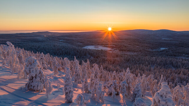 Aerial view of the sun's radiant burst over snow-laden trees, casting long shadows across the winter landscape of Salla, Lapland, Finland.
