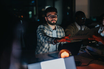 Group of focused business professionals collaborating on a project late at night. The dim lighting sets a serious and intense mood as they work diligently on their laptops and documents. © qunica.com