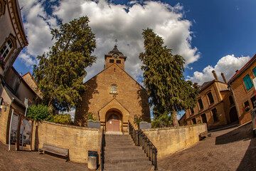 Charming village church in Torgny, Belgium, framed by tall trees under vibrant sky. Quaint stone buildings and cobbled streets enhancing the timeless european atmosphere. 