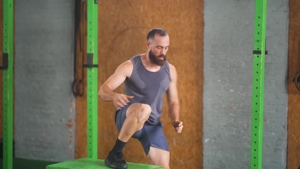 Athletic bearded man performing dynamic box jump exercise in functional training gym with gray tank top and blue shorts - Powered by Adobe