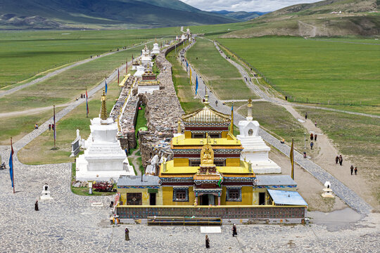 Aerial view of the vibrant gold and white architecture of Bage Mani Stone Wall contrasting against the vast green plains stretching to the distant mountains, Bage Mani Stone Wall, Sichuan, China.