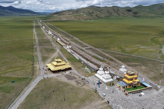 Aerial view of the sprawling Bage Mani Stone Wall and golden-roofed temples amidst a vast, green plateau stretching toward distant mountains, Bage Mani Stone Wall, Sichuan, China.