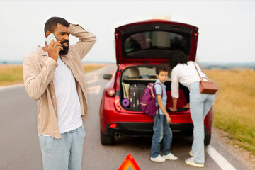Family near broken down car on their way to vacation, man calling on cellphone while woman and boy standing near automobile with open trunk