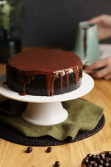 Hand slicing a rich cheesecake with a dark crust, featuring creamy vanilla and chocolate layers, topped with glossy chocolate ganache.