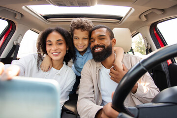 Happy Latin family of three sitting in car, parents and their preteen son taking selfie while traveling by automobile, enjoying road trip adventure