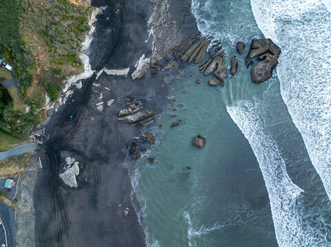Aerial view of the rugged coastline where the dark sands meet the tumultuous turquoise sea, a stark contrast etched by the relentless waves, Port Waikato, Waikato Region, New Zealand.