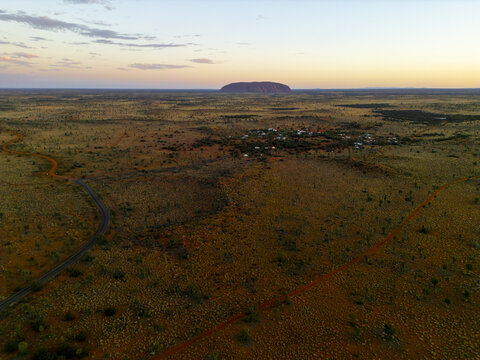 Aerial view of Uluru monolith rising majestically from the flat, arid landscape under a pastel sky, the red earth contrasting with the sparse greenery, Petermann, Northern Territory, Australia.