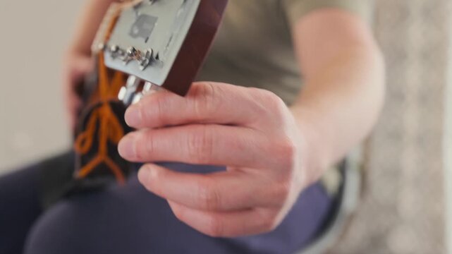 A curly-haired redhead man in glasses tunes his acoustic guitar, focusing on the tuning pegs and strings. Shot at home with soft daylight and a calm atmosphere.