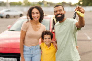 Happy Latin family with child boy posing outdoors next to their brand new automobile, showing key, selective focus, family standing by nice auto on parking spot
