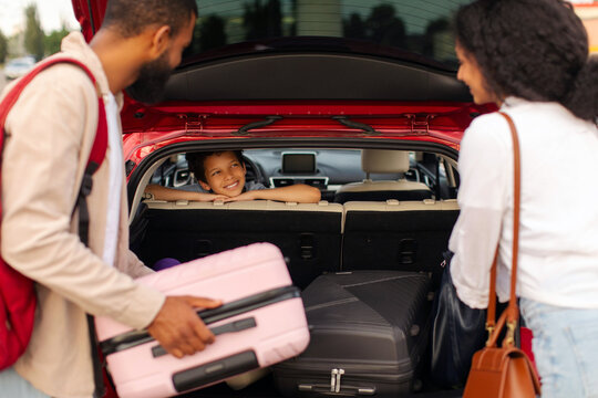Family prepare for travelling by car on holiday, parents putting suitcases in trunk of their vehicle, son sitting inside and looking at them