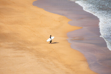 Surfer walking along a sandy beach at sunrise with gentle waves lapping at the shore