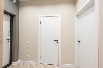 interior shot of a hallway with two white doors. One door has a geometric design, and there is a metal entry door on the left, along with beige walls