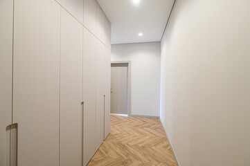 minimalist hallway with light beige wardrobe, white walls, and herringbone wood floor. A door at the end of the hall lets in light