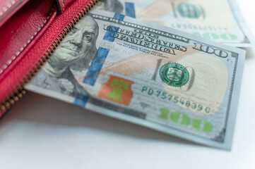 The close-up image shows US$100 bills partially located in a red wallet. The details of the banknotes are visible, including the portrait of Benjamin Franklin.