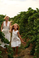 Fototapeta premium Mother and daughter in white dresses running and laughing through a sunny vineyard, enjoying family time, freedom and happiness in nature.