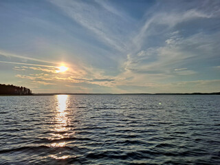 Golden sunset over a peaceful lake with rippling water and a glowing sky. The silhouette of a forest lines the horizon, creating a serene and scenic summer evening atmosphere.