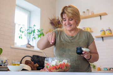 Real middle-aged woman embracing healthy lifestyle, preparing fresh vegetable salad in her home kitchen. Concept of body positivity, self-care, and wellness. menopause and climacteric. Copyspace