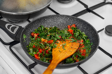 A black frying pan over a gas stove, on which chopped green herb leaves, tomato slices, spices for frying in oil or cooking stews were placed, there is a wooden spatula for stirring