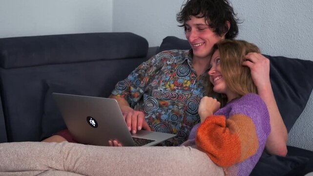 A couple lies on the couch, smiling together as they watch heartwarming videos from trips, reliving bright moments. They laugh as the woman points at the screen, looking at the man with tenderness.