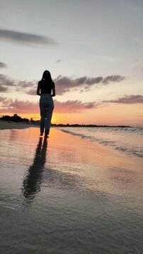 Silhouette of a woman walking barefoot along the shoreline at Batticaloa Beach, Sri Lanka, during a golden sunset, with gentle waves and colorful reflections creating a peaceful coastal scene.
