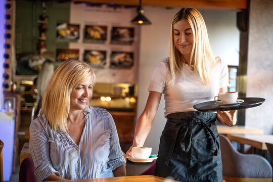 Happy woman, barista and serving customer at cafe for service, payment or order on counter at coffee shop. Female waitress or employee in small business restaurant serving coffee to elder woman - Powered by Adobe