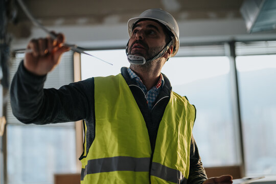A construction worker wearing a helmet and reflective vest inspects wiring at a building site, demonstrating attention to detail and safety measures.