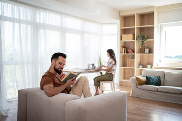 Obraz premium Focused man reading a book while woman works on laptop in living room