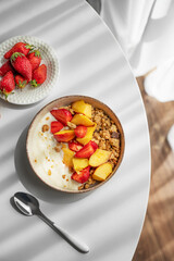 Granola or muesli with natural greek yogurt, strawberry and peach in a bowl on a white table near window