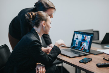 Two professional women collaborate while reviewing a team video conference on a laptop.
