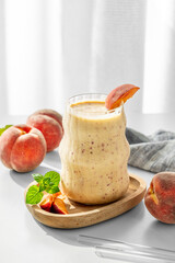 Smoothie with peach and banana in a glass on a wooden board on a white table near window with tropical fruit