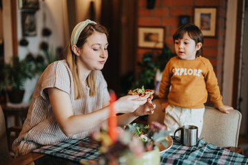 A young mother is seated at a dining table, savoring a slice of pizza, while her toddler attentively observes in a warm and inviting home setting.