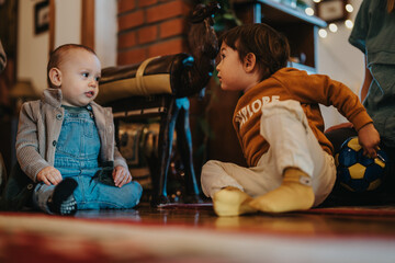 Two children sitting on the floor conversing, one holding a small soccer ball. The bright and warm atmosphere suggests a social interaction or a family setting.