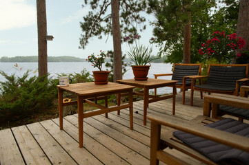 An outdoor wooden table and chairs sit on a deck surrounded by pines, overlooking a shimmering lake – an inviting setting for summer meals and long conversations in cottage country.