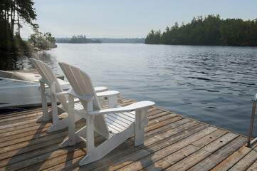 Naklejka premium Two white Adirondack chairs frame a moored boat on a still lake, the soft light of morning illuminating the scene and creating a classic Ontario cottage vibe.