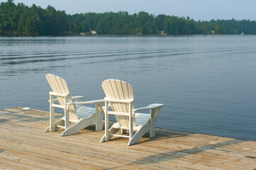 Fototapeta premium Minimalist white Adirondack chairs sit on a dock overlooking a mirror-like lake, the forest horizon glowing in the soft light of early morning – a tranquil retreat in Ontario's Kawarthas.