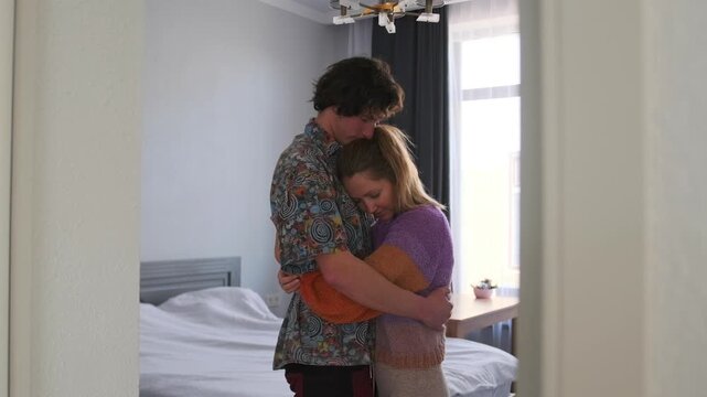 In the bedroom, a young couple stands by the bed, embracing. The woman rests her head on the man chest as he lowers his head and holds her tightly, feeling the depth of their love and connection.