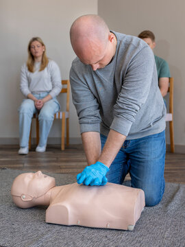 Male student practicing proper chest compression technique on a Basic Life Support (BLS) CPR manikin during a first aid training class, with fellow students watching in the background.