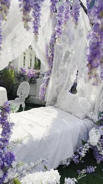A bed with white lace curtain and purple flowers in a outdoor garden, white metal table and chair in the back, paceful place under sunlight