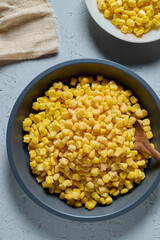 Top view of frozen sweet corn in black metal dish and in white plate with wooden spoon on light gray background.