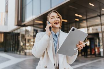Confident woman wearing formal attire conversing on the phone while holding a tablet, standing in front of a modern building, showcasing professional and dynamic energy