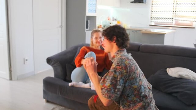 The man kneels in front of the woman sitting on the couch, slips a ring onto her finger, and gently kisses her hand. She smiles, touched by the moment, and embraces him, sharing the joy and excitement