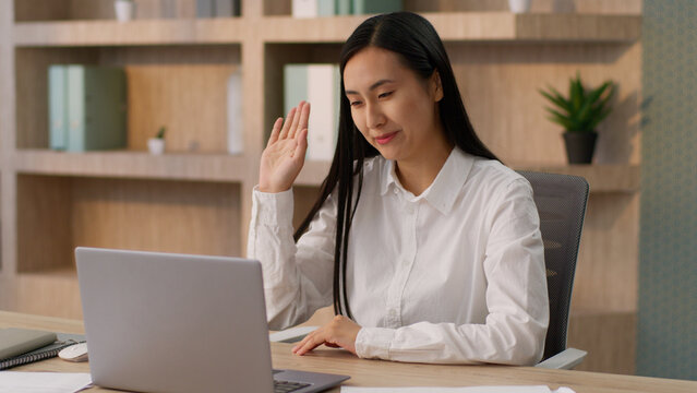 Happy laughing Asian Korean Chinese woman female worker Japanese girl boss businesswoman talking laptop pc waving hand hello gesture video call online conference computer business meeting in office
